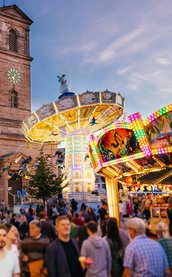 Menschen genießen die Michaelis-Kirchweih Fürth mit beleuchteten Fahrgeschäften vor der Stadtkirche.