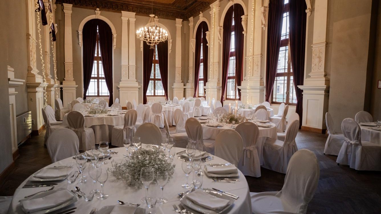 Saal im Logenhaus Fürth mit rund gedeckten weißen Tischen, Kerzen, Stuhlhussen, großen Fenstern und Kronleuchtern