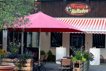 Außenansicht von Frankies Hofladen mit Sonnenschirm, Sitzgelegenheiten, Blumen und Stehtischen am Eingang