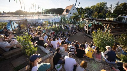 Menschen sitzen auf halbrunden Gartenrängen zwischen Pflanzen und warten im Sonnenschein auf den Auftritt.
