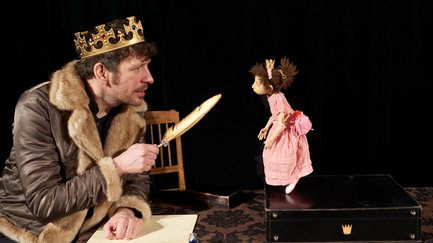 Mann mit Krone und Feder am Tisch; vor ihm ein kleiner Koffer mit Puppe beim internationalen figuren.theater.festival Fürth.