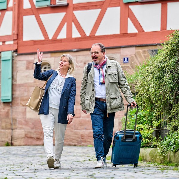 Zwei Best-Ager-Touristen mit Koffern laufen über den Kirchplatz in Fürth, hinter ihnen ein Fachwerkhaus.