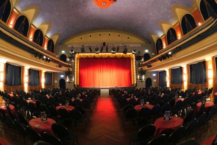 Saal der Comödie Fürth mit Tischen und Stühlen, roter Vorhang vor der beleuchteten Bühne