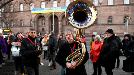 Kleine Bläsergruppe mit Sousaphon, Trompete und Trommler spielt im Freien vor umstehenden Zuschauern.