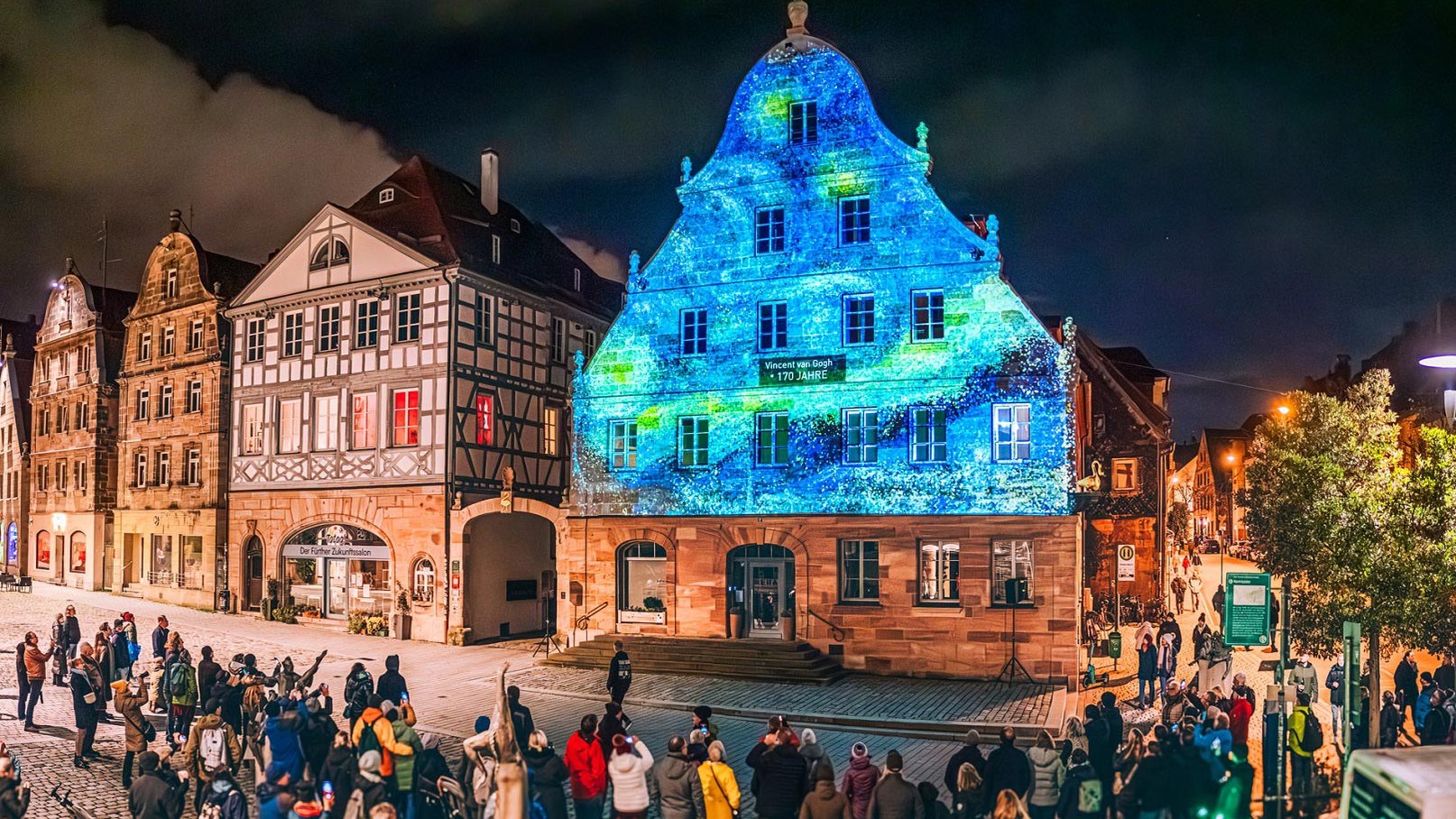 Bunt beleuchtete Hausfassade am Grünen Markt in Fürth, umgeben von vielen Menschen in der Altstadt.