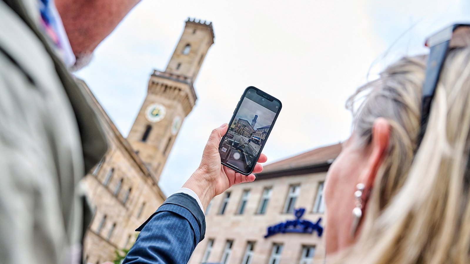 Frau hält Handy und fotografiert das Fürther Rathaus, Mann steht daneben, Rathaus im Hintergrund unscharf