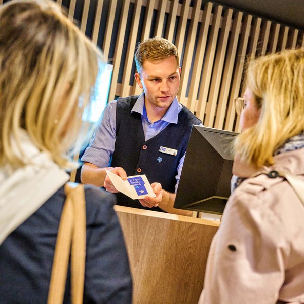 Ein Hotelmitarbeiter an der Rezeption übergibt zwei Gästen von hinten gesehen eine Zimmerkarte.
