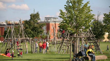 Kinder klettern auf großen Holzgerüsten auf einer Wiese, während Eltern im Park daneben sitzen.