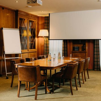 Gemütlicher Tagungsraum mit Holztisch, sieben Stühlen, Flipchart, Leinwand, Teppichboden und warmen Farben