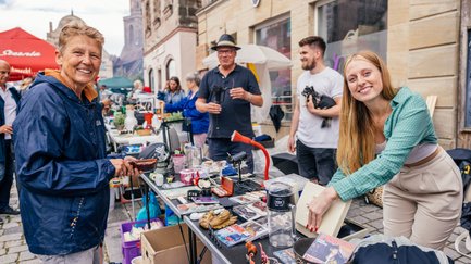 Käufer und Verkäuferin lächeln an einem Flohmarktstand auf dem Graffelmarkt, umgeben von weiteren Ständen.