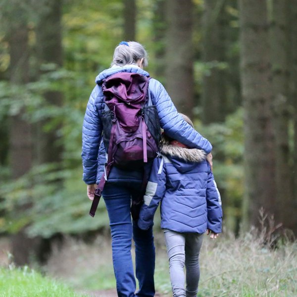 Zwei Kinder toben auf einem Waldweg, dahinter geht eine Mutter mit ihrem Arm um ein weiteres Kind.