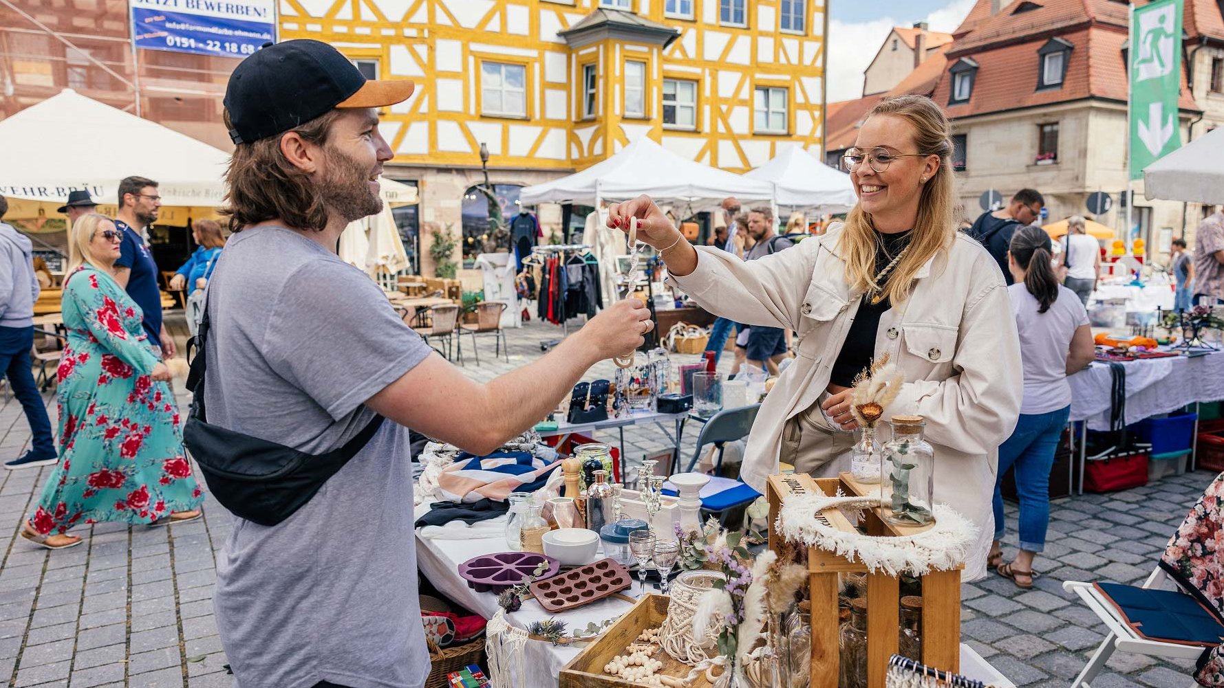 Flohmarktstände in der Altstadt; eine Verkäuferin reicht einem Kunden ein gekauftes Stück am Stand.