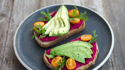 Zwei reich belegte Brote mit Rote-Bete-Aufstrich, Avocadoscheiben und Kirschtomaten auf Teller.