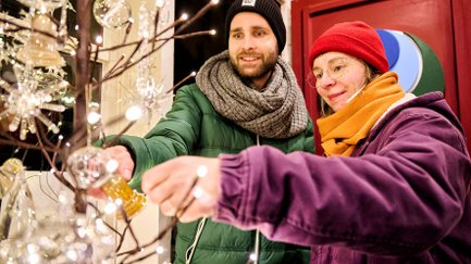 Ein Paar steht im Glasbläser-Häuschen und betrachtet eine gelbe Glasglocke am geschmückten Bäumchen.
