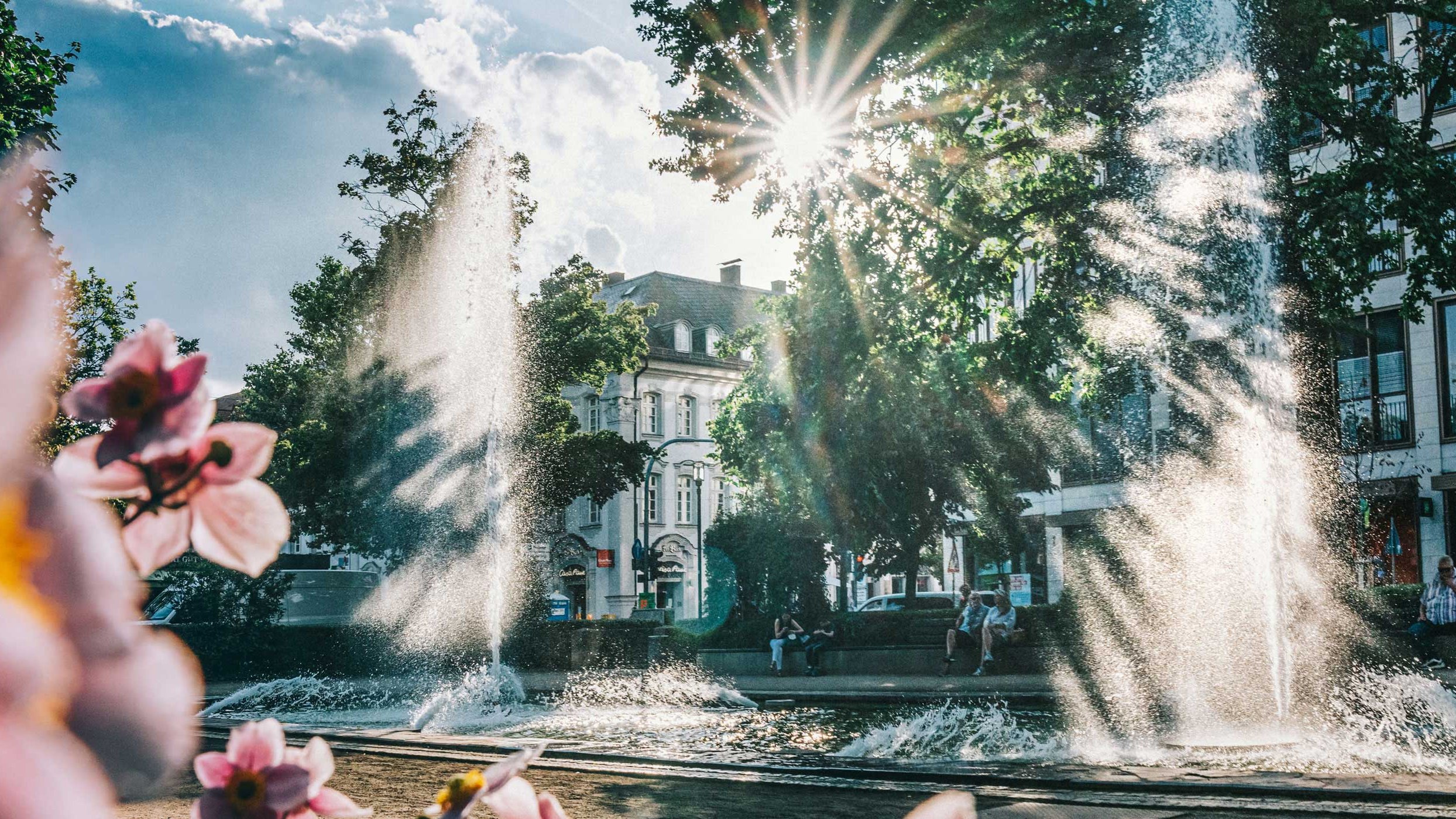 Springbrunnen auf der Fürther Freiheit, umgeben von Bänken mit sitzenden Menschen und grünen Bäumen.