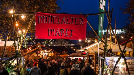 Rotes Banner mit „Mittelaltermarkt“ markiert den Eingang, unter dem Besucher in das bunte Treiben gehen.