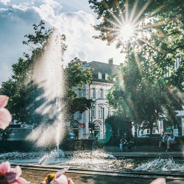 Springbrunnen auf der Fürther Freiheit, umgeben von Bänken mit sitzenden Menschen und grünen Bäumen.