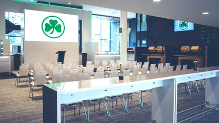 Blick auf Bühne mit Logo auf der Leinwand, davor Stuhlreihen und Stehtische für Gäste