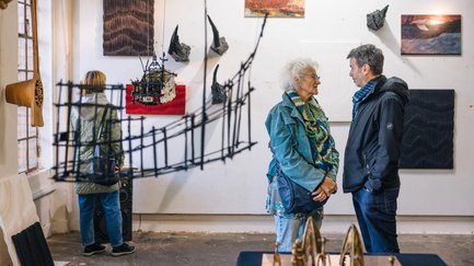 Drei Personen betrachten im Gastspiel moderne Kunstwerke an Wand, Tisch und Decke in hellem Ausstellungsraum.