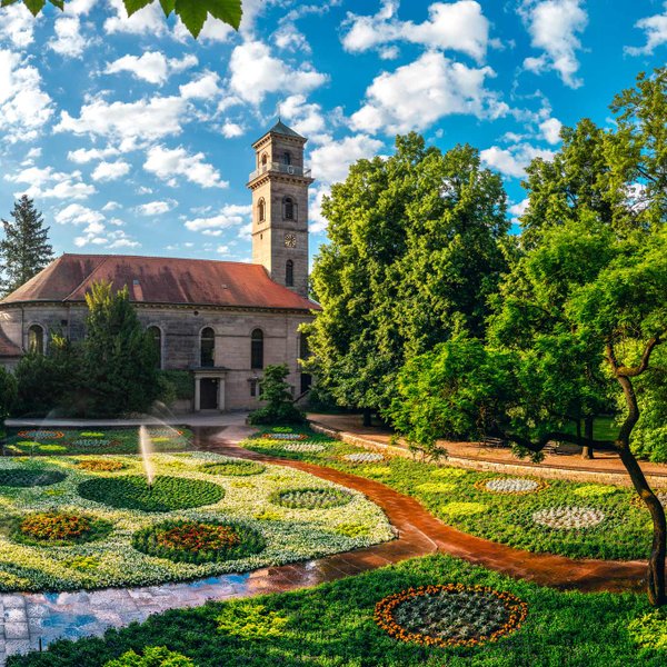 Blick über die farbenfrohen, kreisförmigen Blumenbeete des Empfangsgartens im Fürther Stadtpark auf die historische Auferstehungskirche bei strahlendem Sonnenschein.