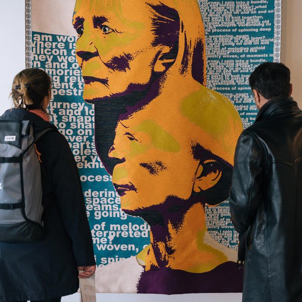 Zwei Personen stehen vor blauem Wandteppich mit gelb dominierten Porträtmotiven und weißem Schriftzug.