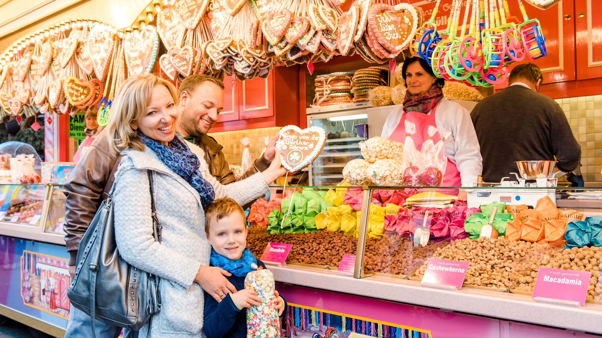 Familie steht an einem Süßwarenstand der Kirchweih mit gebrannten Nüssen, Popcorn und Lebkuchenherzen.