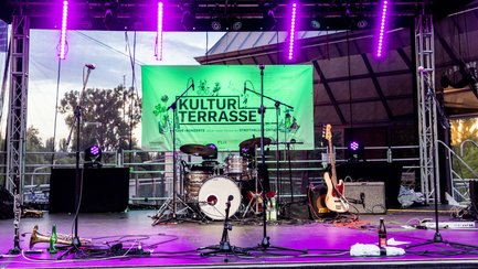 Beleuchtete Bühne mit Musikinstrumenten, Mikrofonen und grünem Kulturterrasse-Banner im Hintergrund.