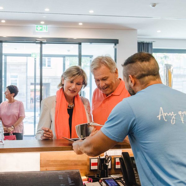 Ein Paar lässt sich am Empfangstresen eines Hotels von einem Mitarbeiter in blauem Shirt beraten.