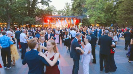 Paare in eleganter Abendkleidung tanzen auf der Tanzfläche im Stadtpark zur Live-Musik einer Band, umgeben von Bäumen.