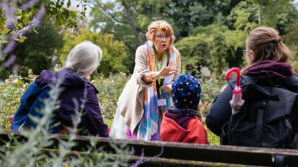 Gruppe sitzt im Rosengarten und lauscht einer Stadt(ver)führerin während einer geführten Tour
