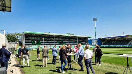 Personen an Stehtischen am Spielfeldrand, im Hintergrund die Nordtribüne des Sportparks