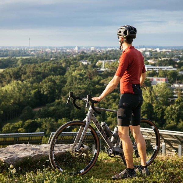 Ein Radfahrer steht mit seinem Rad auf einem Hügel und blickt über grüne Felder auf die Stadt.