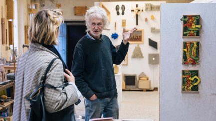 Atelierbesitzer erklärt einer Besucherin drei farbige Kunstwerke, umgeben von Werkbänken und weiteren Kunstwerken.