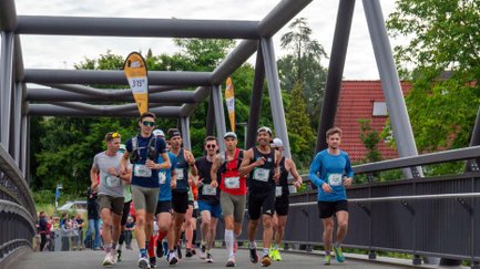 Läufergruppe mit Startnummern überquert eine Brücke, Zuschauer klatschen und feuern sie am Streckenrand an