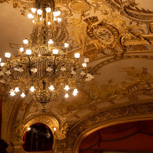 Kronleuchter im Stadttheater hängt vor bemalter Decke mit Stadtwappen und Engelsfiguren.