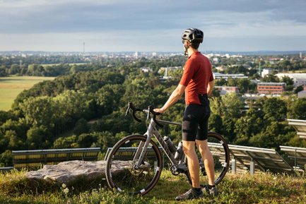 Ein Radfahrer steht mit seinem Rad auf einem Hügel und blickt über grüne Felder auf die Stadt.