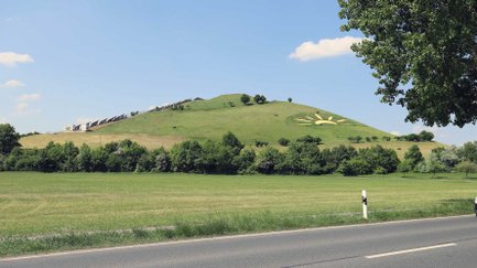 Ein begrünter Hügel mit Solaranlagen und einem großen Sonnen-Symbol liegt hinter einer Landstraße.