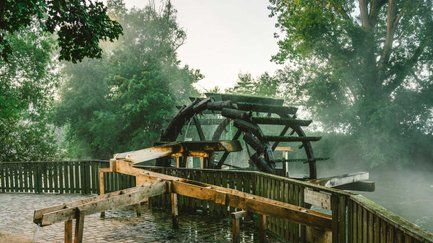 Ein historisches Schöpfwasserrad aus Holz dreht sich an einem Flussufer im morgendlichen Nebel.