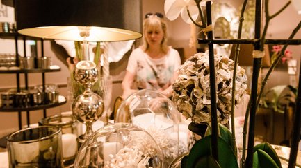 Frau schaut Wohnaccessoires und Kleinmöbel in einem Geschäft in Fürth an, leuchtende Lampe und Kunstblume im Vordergrund