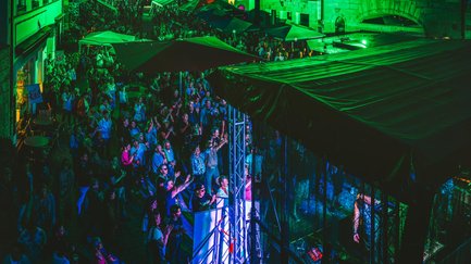 Besucher feiern und schlendern zwischen Bühne und Ständen auf dem grünen, beleuchteten Markt.