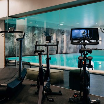 Fitnessstudio im Hotel Forsthaus Fürth mit Laufband und Biketrainer vor Glaswand zum Schwimmbad