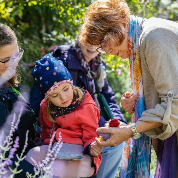 Märchenerzählerin zeigt einem Mädchen auf dem Schoß der Mutter eine Puppe während eines Stadtspaziergangs.