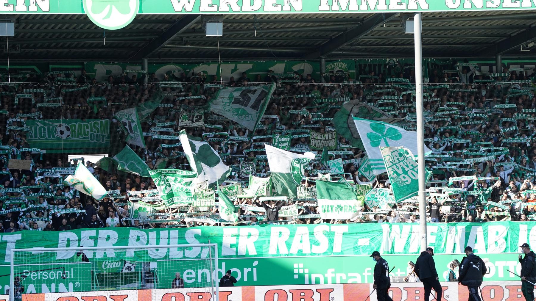Greenkeeper bereiten den Rasen im Sportpark Ronhof vor, während Fans mit Schals und Fahnen die Nordtribüne füllen.