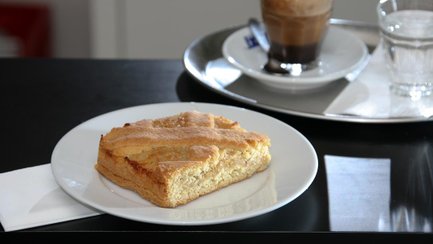 Ein Stück Zitronenkuchen auf einem weißen Teller mit Servierte, im Hintergrund ein Tablett Kaffee.