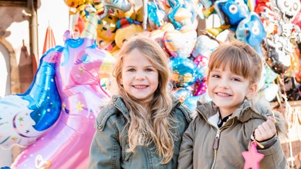 Zwei lächelnde Kinder stehen vor einem bunten Luftballonstand mit vielfältigen Motiven und Formen.