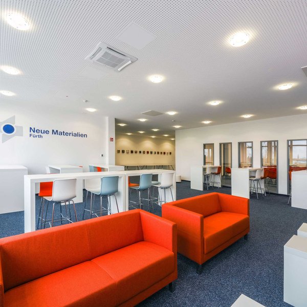 Großer Raum bei Neue Materialien Fürth mit Stehtischen, hohen Stühlen, Tageslicht und Logo an der Wand