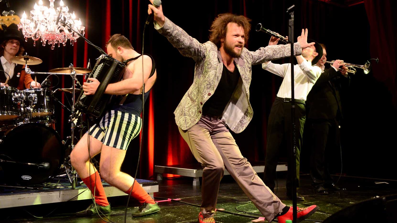 Band spielt live auf der Bühne, Sänger tanzt im Vordergrund, Schlagzeug, Akkordeon und Trompeten begleiten.