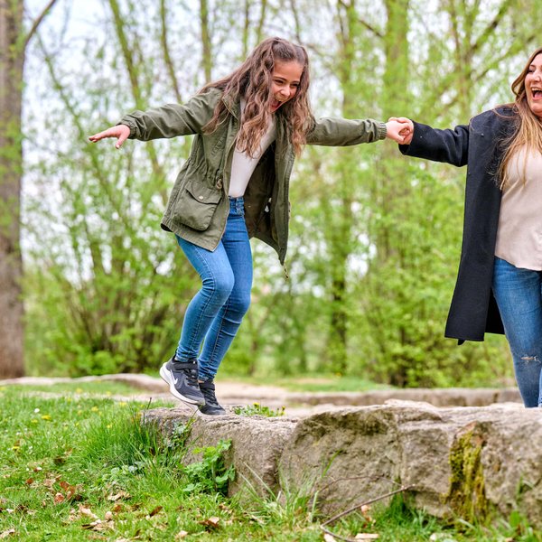 Mutter und Tochter balancieren lachend an den Händen haltend über große Steine auf einer Wiese.