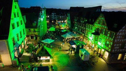 Historische Gebäude am Grünen Markt leuchten grün, Besucher sitzen an Bierbänken und Ständen.