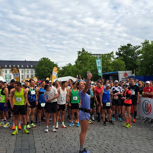 Frau macht Selfie vor Marathonstart, Teilnehmer in Sportkleidung und Zuschauer im Hintergrund